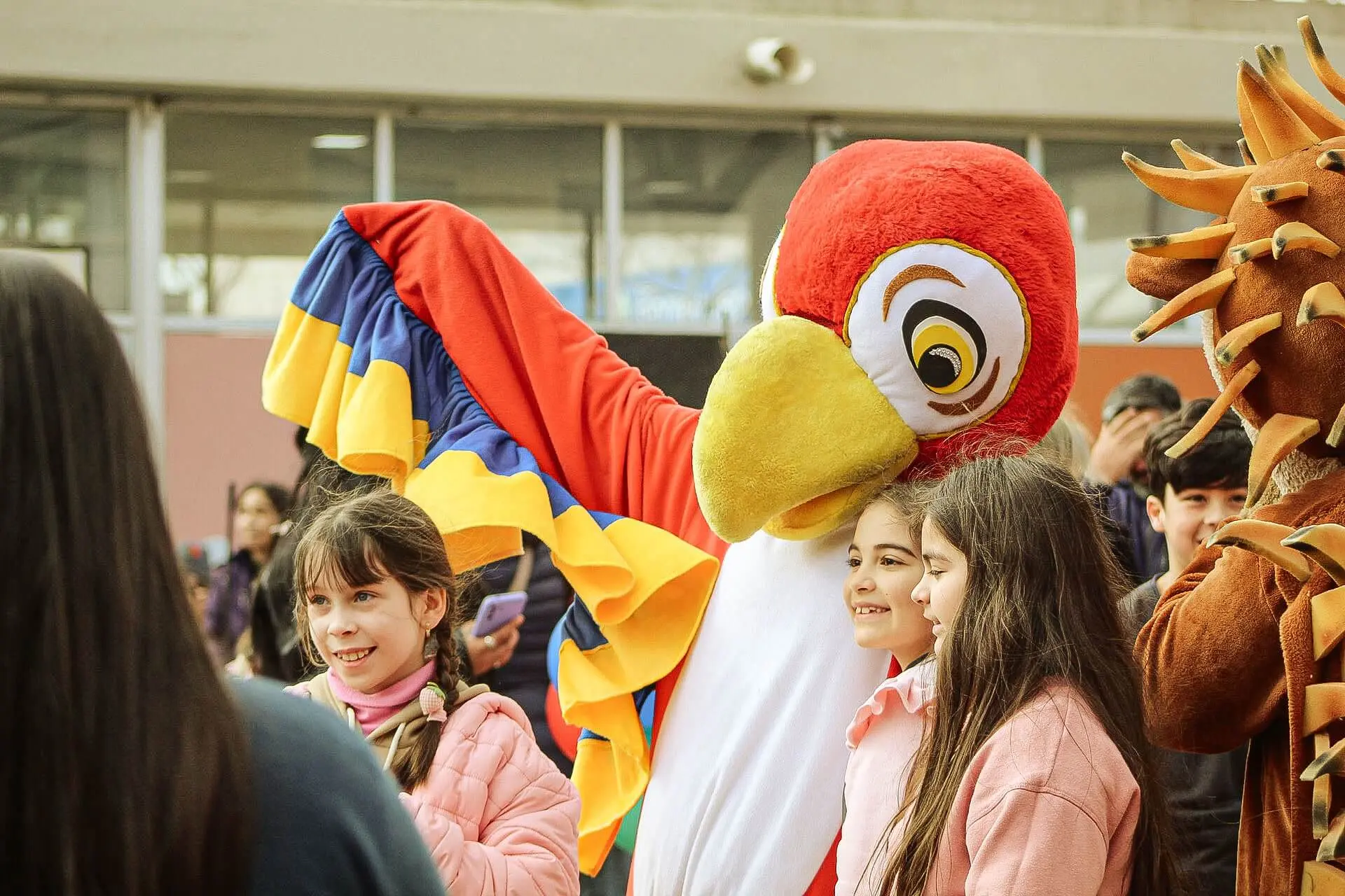 Niñas sonríen junto a una persona disfrazada de loro de colores.