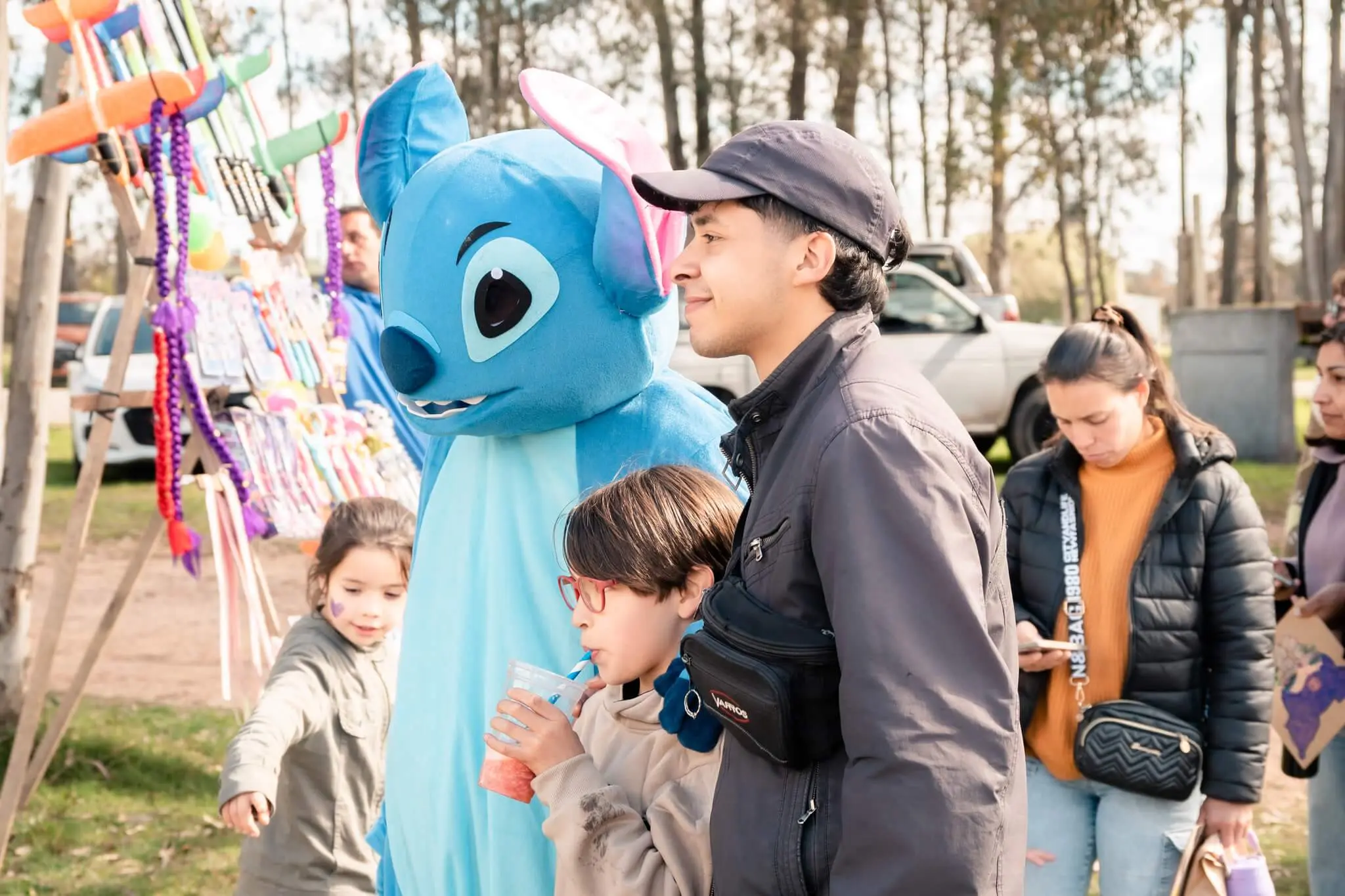 Un niño y su padre se ríen junto a una persona disfrazada de Stitch, entre puestos de globos.
