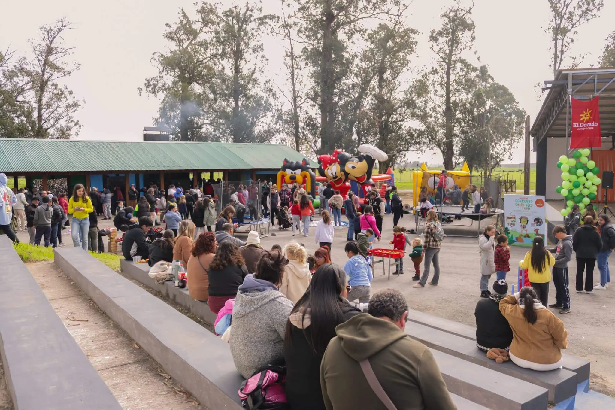 Familias en las gradas del parque frente a los juegos y puestos de la fiesta.
