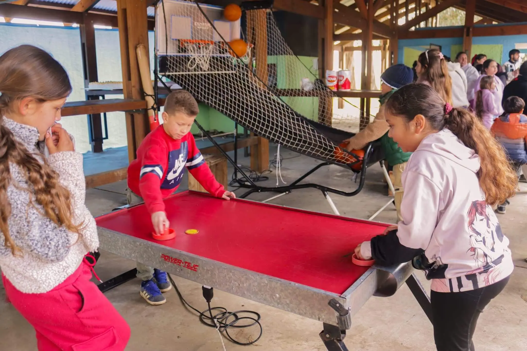 Niños juegan al tejo de mesa bajo un techo de madera, con un juego de básquet al fondo.