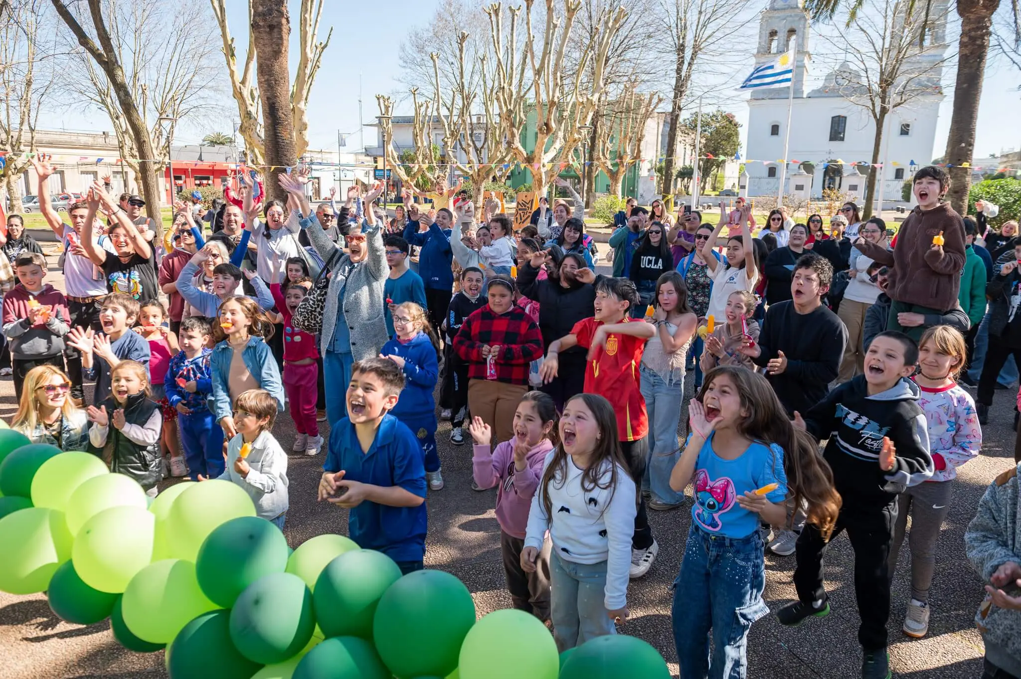 Decenas de niños saltan y festejan con los brazos en alto en la plaza, frente a la iglesia y detrás de una fila de globos verdes.
