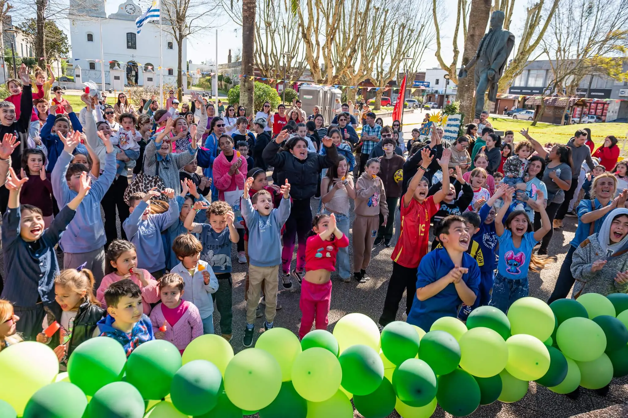 Niños y familias bailan con las manos arriba frente al escenario, con la iglesia y la bandera uruguaya al fondo.