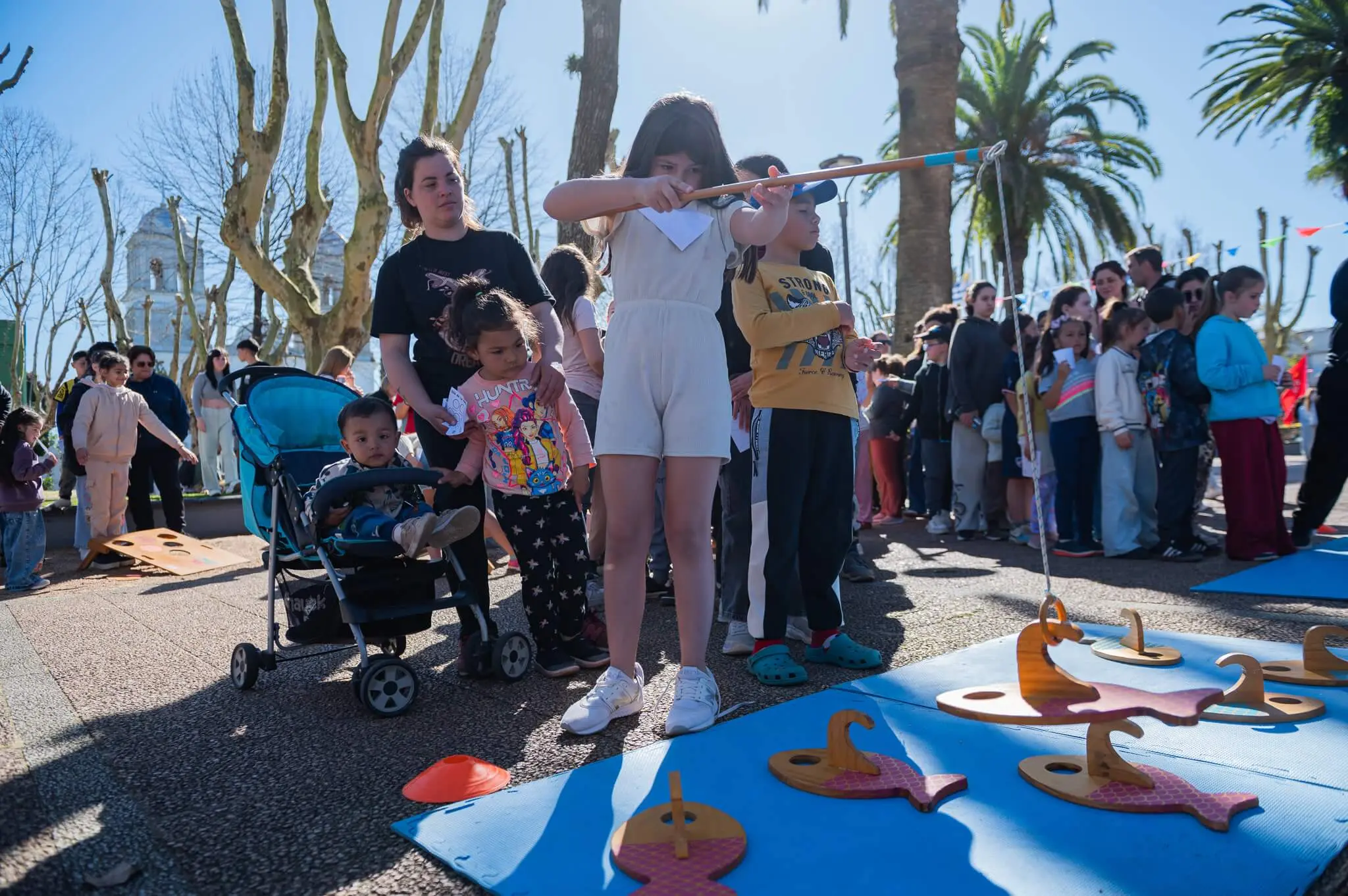 Una niña juega a pescar figuras de madera con una caña mientras otros niños esperan su turno en la plaza.
