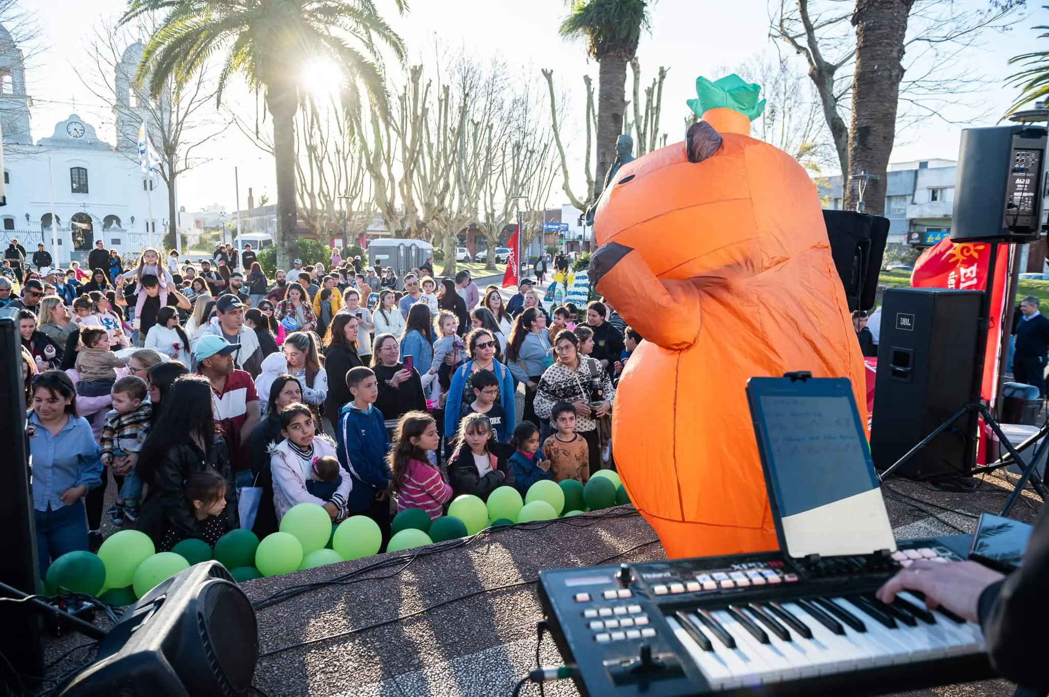 Una capibara inflable gigante junto al escenario, frente a la multitud de niños y familias en la plaza.