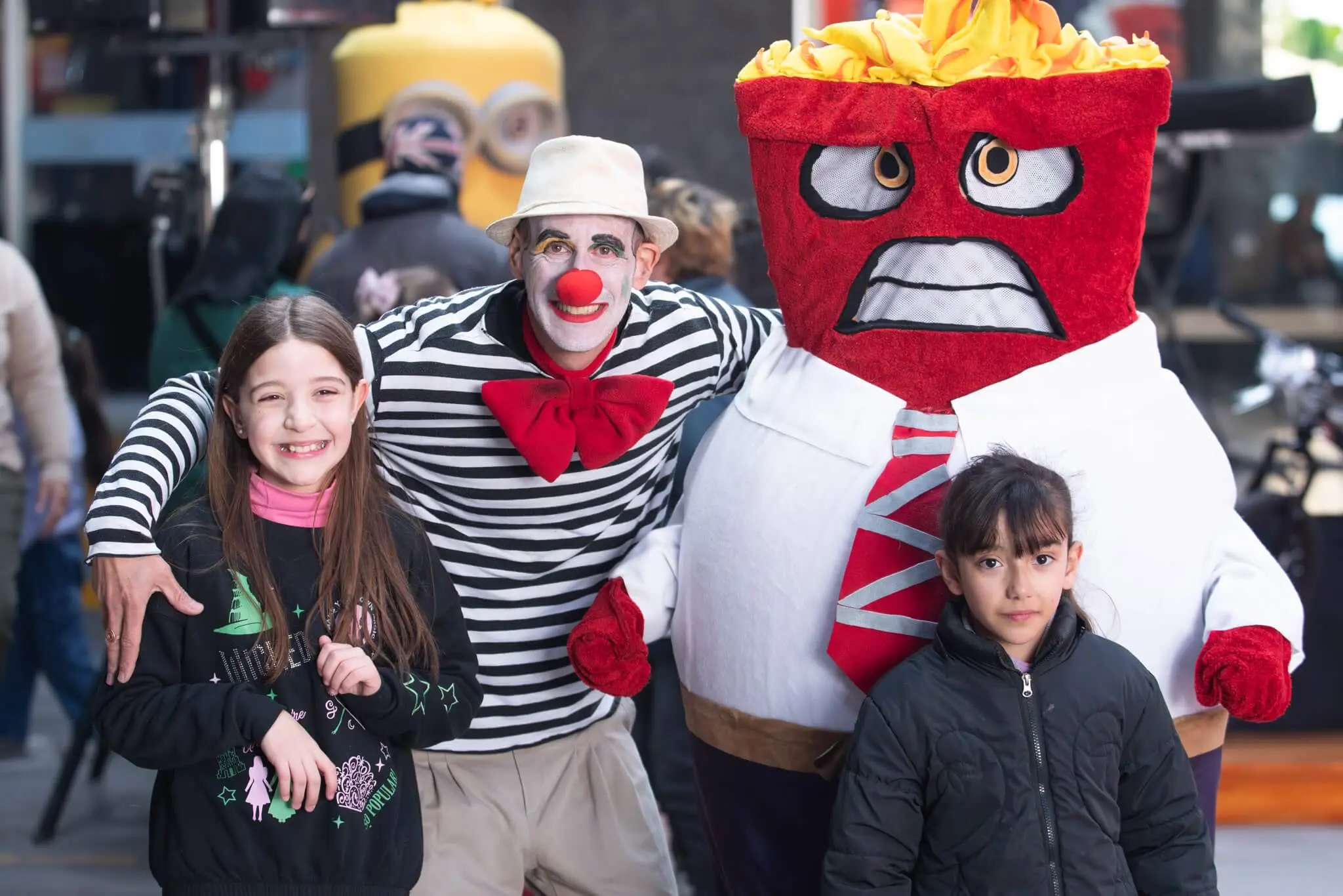 Dos niñas posan sonrientes con un payaso y una botarga del personaje Furia, de Intensamente.